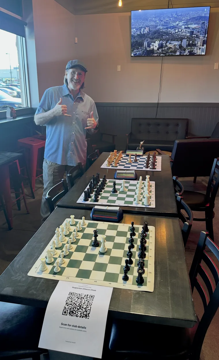 Chuck Price standing at the head of a long table in the window-lit room at The Stable Bar & Grill, three chess sets laid out and ready along it, giving a two-thumbs-up to the camera.