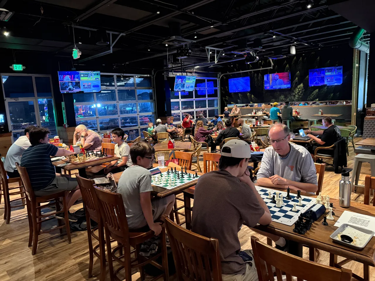 A wide view of a club night in progress: several chess boards on wooden tables with players at each, more people eating and talking at tables beyond, under a high dark ceiling and a row of screens.